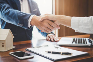 Two professionals shake hands over a desk after agreeing on a deal; a small house model, laptop, and paperwork are visible on the table.