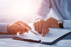 Two people sign a document at a desk, one holding a pen and the other pointing to the text of an agreement.